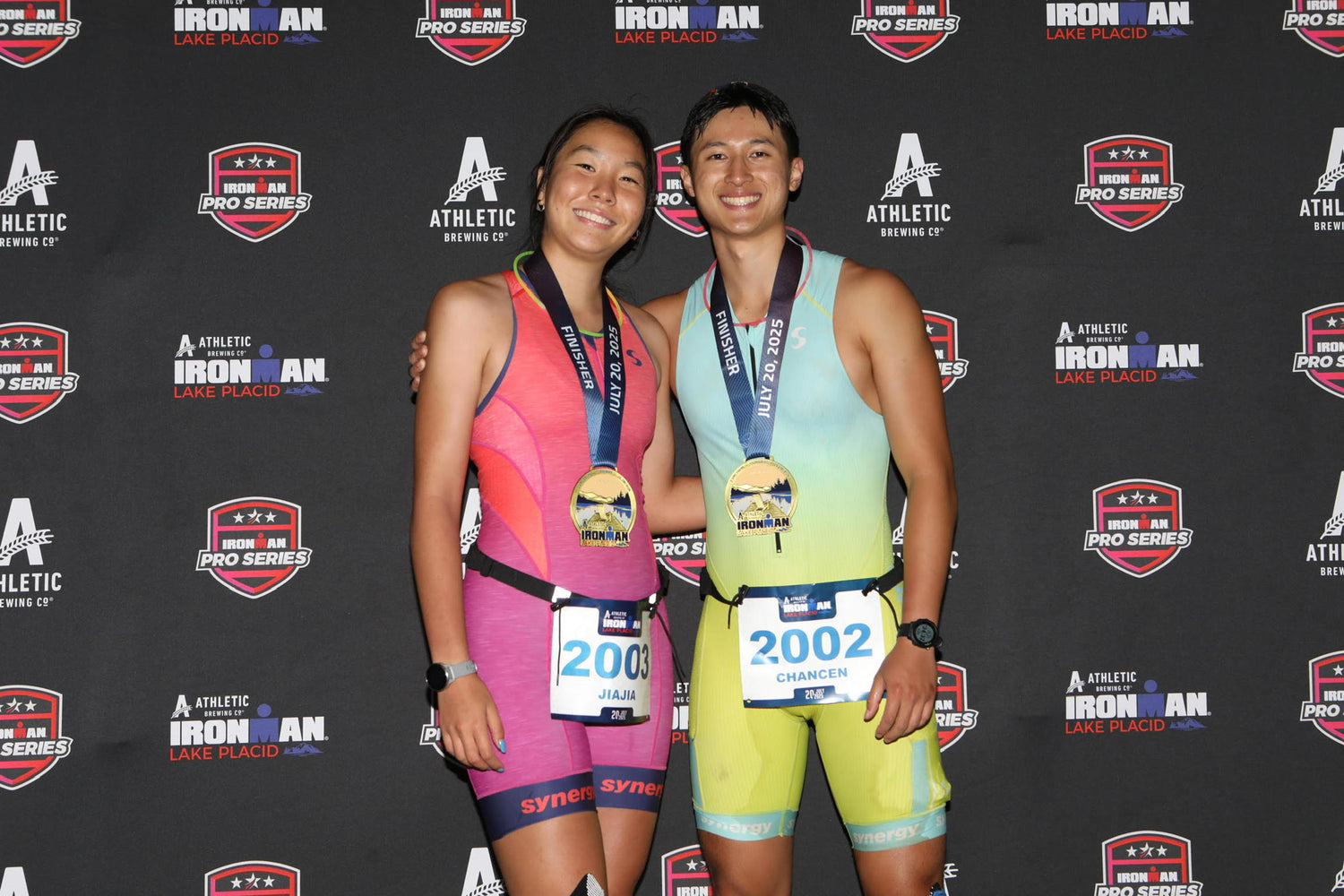 Two athletes standing together with medals at an Ironman event, posing in front of a branded backdrop.